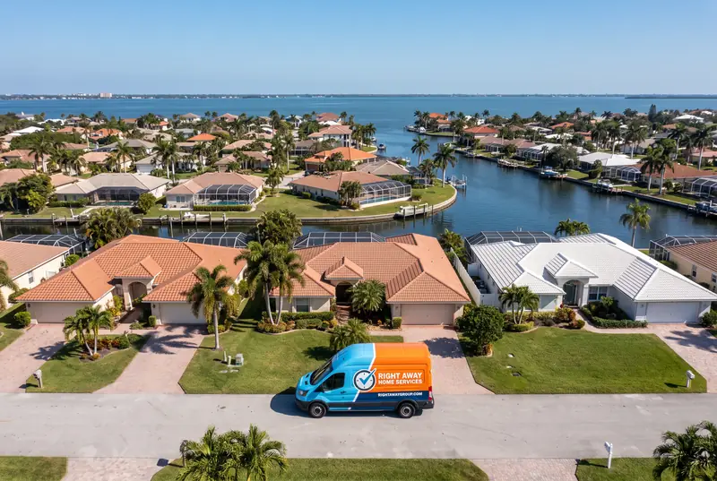 An aerial view of a bayside suburb with tile roofs, screened pools, palms and a canal running to the bay, a single sky-blue and orange service van on the street below